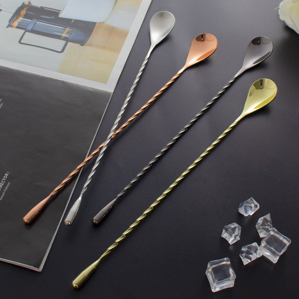 Four long bar spoons with twisted handles and teardrop-shaped heads in silver, rose gold, black, and gold finishes, arranged on a dark surface with a magazine and clear ice cubes as props.