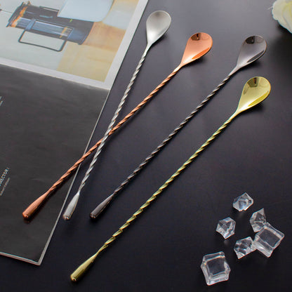 Four long bar spoons with twisted handles and teardrop-shaped heads in silver, rose gold, black, and gold finishes, arranged on a dark surface with a magazine and clear ice cubes as props.