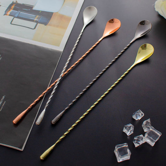 Four long bar spoons with twisted handles and teardrop-shaped heads in silver, rose gold, black, and gold finishes, arranged on a dark surface with a magazine and clear ice cubes as props.